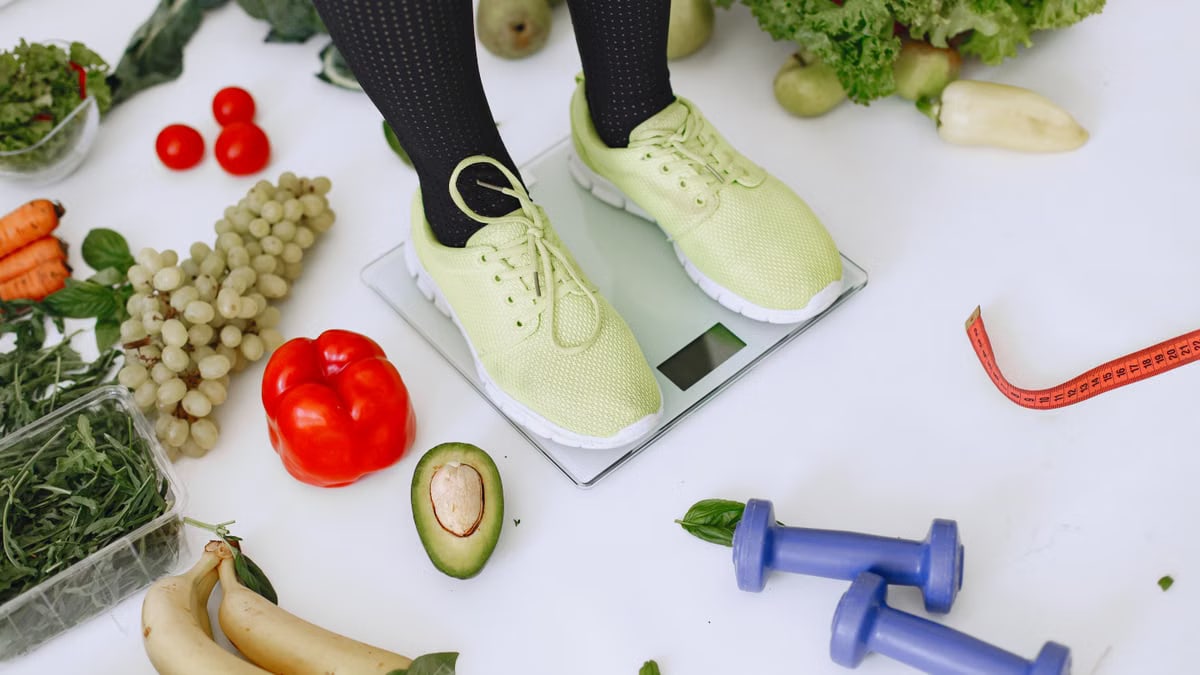 Person standing on scales surrounded by fresh produce and fitness equipment symbolizing a balanced lifestyle.